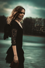 Portrait of a beautiful girl with black wings in the form of a black angel standing on the river bank that is covered with ice and sky with clouds