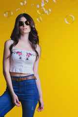 Portrait of beautiful young brunette with glasses in white top and jeans standing on a yellow background in the Studio .Fashion looks elegance clothes..bubbles