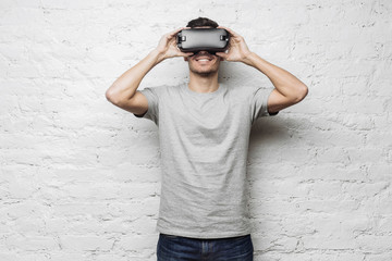 Technology, gaming and people concept. Young attractive caucasian man wearing grey t-shirt and virtual reality headset or 3d glasses, playing video game or watching movie against white brick wall