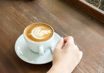 Start The Day with Good Cup of Coffee Concept. Perspective View Business Asian Woman Hand Holding Cup of Cappuccino Coffee with Tree Design Foam on Wooden Panel Table by Window of Nature Garden View
