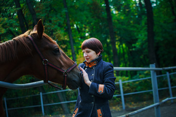Obraz premium A woman in a blue long coat and with short dark brown hair extended her hand to stroke the horse. The horse pricked up his ears. A train for horses against the background of a green forest.