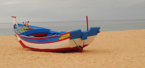 Fototapeta premium Barco solitário na praia, parado na areia com horizonte por trás, cores brancas, azuis e vermelhas