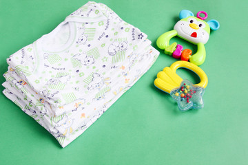 concept of extending family, preparing and expecting for baby: stack of ironed newborn baby clothes with colorful rattles on green background with copy space