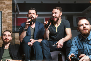 A company of four men with a beard sitting on the couch at home with beer and chips, two of them with joysticks in hand playing computer video games, the other carefully watching the game. 