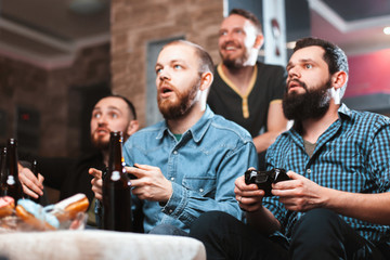 A company of four men with a beard sitting on the couch at home with beer and chips, two of them with joysticks in hand playing computer video games, the other carefully watching the game. 