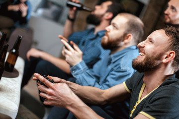 A company of four men with a beard sitting on the couch at home with beer and chips, two of them with joysticks in hand playing computer video games, the other carefully watching the game. 