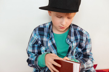 Fototapeta na wymiar Childhood and leisure. Image of schoolboy in stylish checked shirt and cap opening red book isolated over white background wanting to read interesting stories and fairy tales