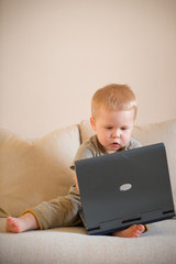 Cute little toddler boy sitting on the sofa with his laptop and looking at the window. Learning with computer. Early development. Child and computer.