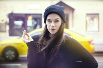 Young pretty woman smoking outside. Hipster outfit, wearing black hat and t-shirt, city yellow taxi...