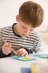 Portrait of little school boy sitting at the table and drawing with yellow paint. After school activities at home. Education. Small boy creating handmade cards.