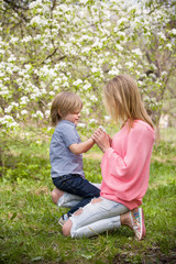 Fototapeta premium Beautiful blond woman with her little adorable kid boy in the blooming park on a sunny spring day. Happy mother with her son sitting on the green grass and enjoying flowers. Mum and child outdoors