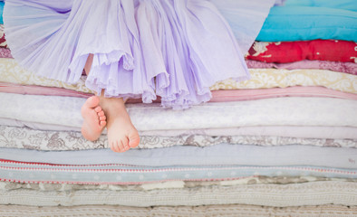 The fairy tale the princess on a pea. Little girl in lilac tatu skirt sitting on the high bed. Bare foot if the girl. Legs of a little girl sitting on a pile of colorful mattresses.