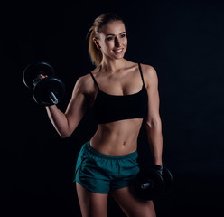 Cute athletic model girl in sportswear with dumbbells in studio against black background. Ideal female sports figure. Fitness woman with perfect sculpted muscular and tight body.