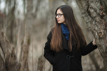 Young beautiful girl in a black coat blue scarf glasses walking in the autumn / spring forest park. An elegant brunette girl with gorgeous extra long hair. Vintage film effect saturated colors.