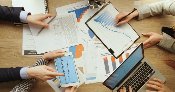 Business team are checking financial charts and graphs, pointing and working with tablet and laptop, writing in notebook at office wooden desk. Hands top view. Quick motion. Time lapse. Team work