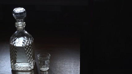 A bottle of vodka and a glass on a wooden table. Dark background