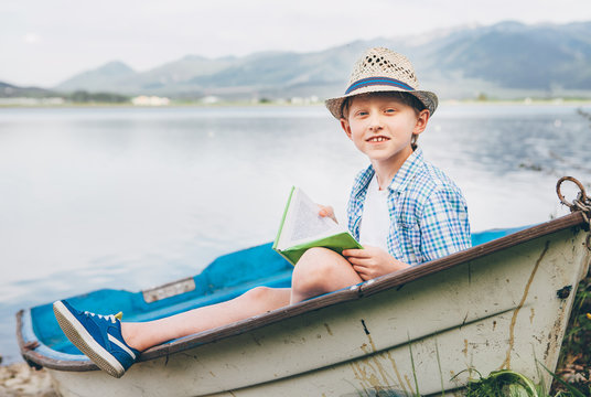 Boy with book sits in old boat on the mountain lake bank