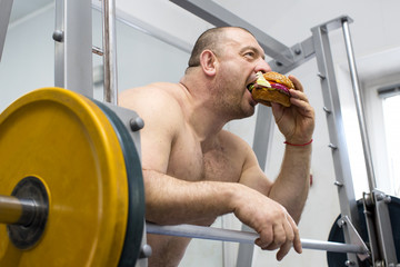 A big fat hungry man eats a hamburger with meat and cheese in the gym