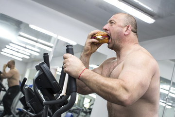 A big fat hungry man eats a hamburger with meat and cheese in the gym