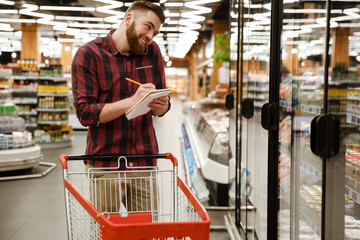 Cheerful young man in supermarket talking by phone writing notes