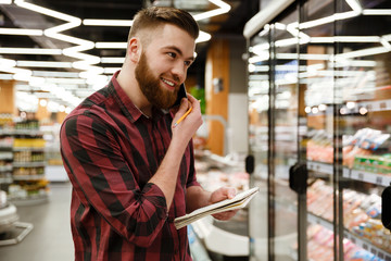 Happy young man standing in supermarket talking by phone