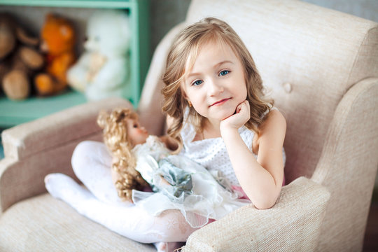 Cute blond girl in white dress sits in chair With a doll in hands
