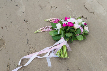 Delicate bridal bouquet with white and pink peony and greenery with long ribbon lying on wet sand on the bank of river