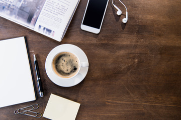 Top view of coffee cup, smartphone with blank screen and earphones, notebook with pen and newspaper on wooden table