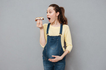 Pregnant happy lady eating eclair. Looking aside.