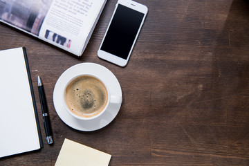 Top view of coffee cup, smartphone with blank screen, notebook with pen and newspaper on wooden table