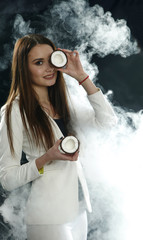 Young girl in a white jacket holds a coconut near her eye and smiles on a black background, covered with smoke (vapor)