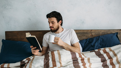 Bearded man reading a big book lying in his bedroom.