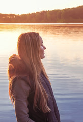 Beautiful girl standing near water outdoors. Cute Long-haired blonde sunset lake. Young woman thinking about something river during golden
