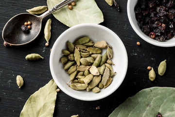 Cardamon seeds in a white ceramic bowl among other condiments on a black wooden table, top view