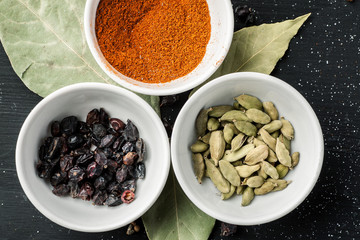 Paprika powder, barberry and cardamon seeds in white ceramic bowls on a black wooden table, top view