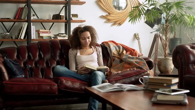 young happy Latin woman read interesting paper book for education sitting alone on comfort leather sofa in the cozy living room at home with bookshelf near window during sunny day
