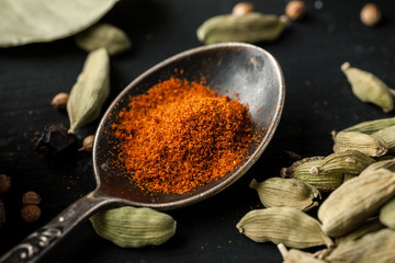 Ground paprika in a metal spoon with cardamon seeds around on a black wooden table, selective focus