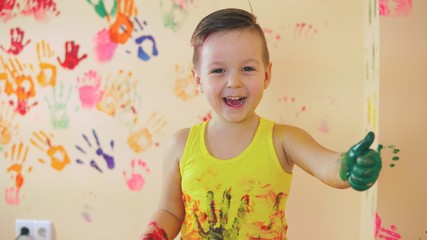 Close up veiw of cute little boy with painted hands after leaving his colorful handprints on the wall and showing his thumb up. Young happy family. Mother and child concept