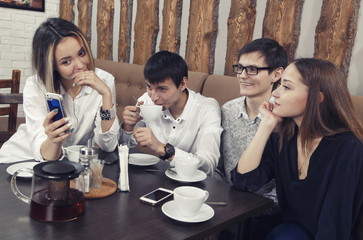 Group of young people from two couples of guys and girls have a tea time in the cafe and looking on the smartphone absorbedly