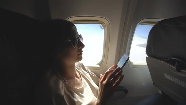Tourist woman sitting near airplane window at sunset and using mobile phone during flight