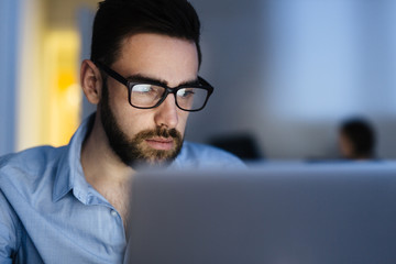 Portrait of handsome bearded man wearing glasses working with laptop in dark office late at night,...