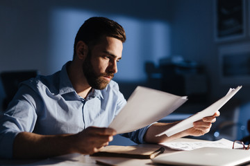 Portrait of focused bearded businessman wearing casual clothes working with documentation in dark room late at night, his face lit by lamp light - obrazy, fototapety, plakaty