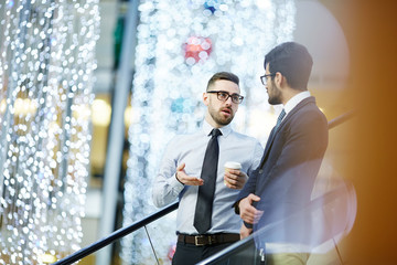 Businessman with drink explaining idea to colleague while moving down on escalator