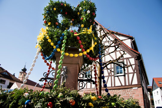 Osterbrunnen geschmückt mit aufgehängten Eiern vor Fachwerkhaus und blauem Himmel