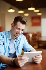 Portrait of modern young man playing video game using portable digital tablet in cafe and smiling joyfully