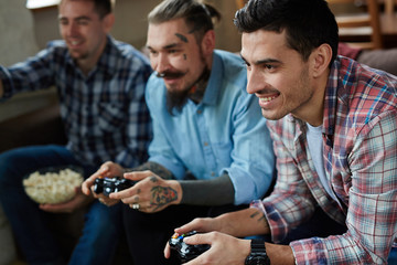 Portrait of three excited adult men enjoying video game competition and smiling cheerfully holding wireless controllers while sitting on couch in living room with popcorn