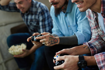 Young men with tattoos enjoying video game competition and smiling cheerfully holding wireless controllers while sitting on couch in living room with popcorn