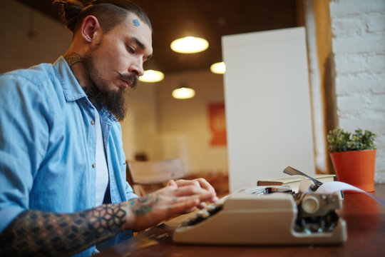 Side view portrait of creative tattooed man using old-school vintage typewriter on wooden table in cafe, typing story looking for inspiration