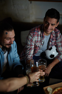 High Angle Portrait Of Group Of Friends Meeting At Home, Sitting On Couch In Dark Room Toasting With Beer And Eating Pizza While Watching Football Match On TV
