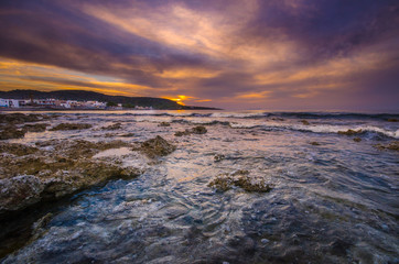 Gorgeus sunset over sea with waves, rocks and traditional greek village of Milatos, Crete, Greece.